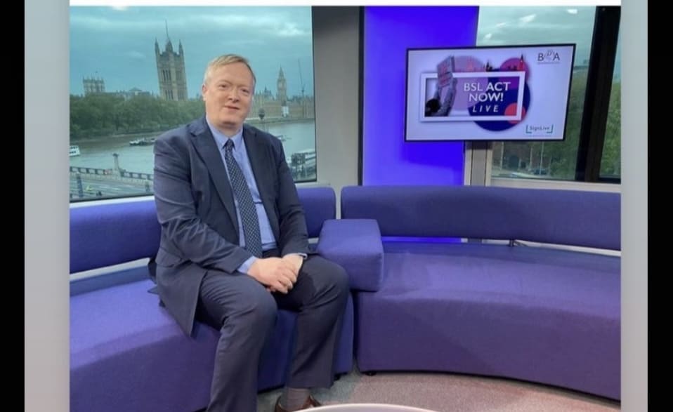 A man in a suit sitting on a purple couch in a tv studio, with a view of a cityscape through the window and a tv screen displaying news graphics behind him.