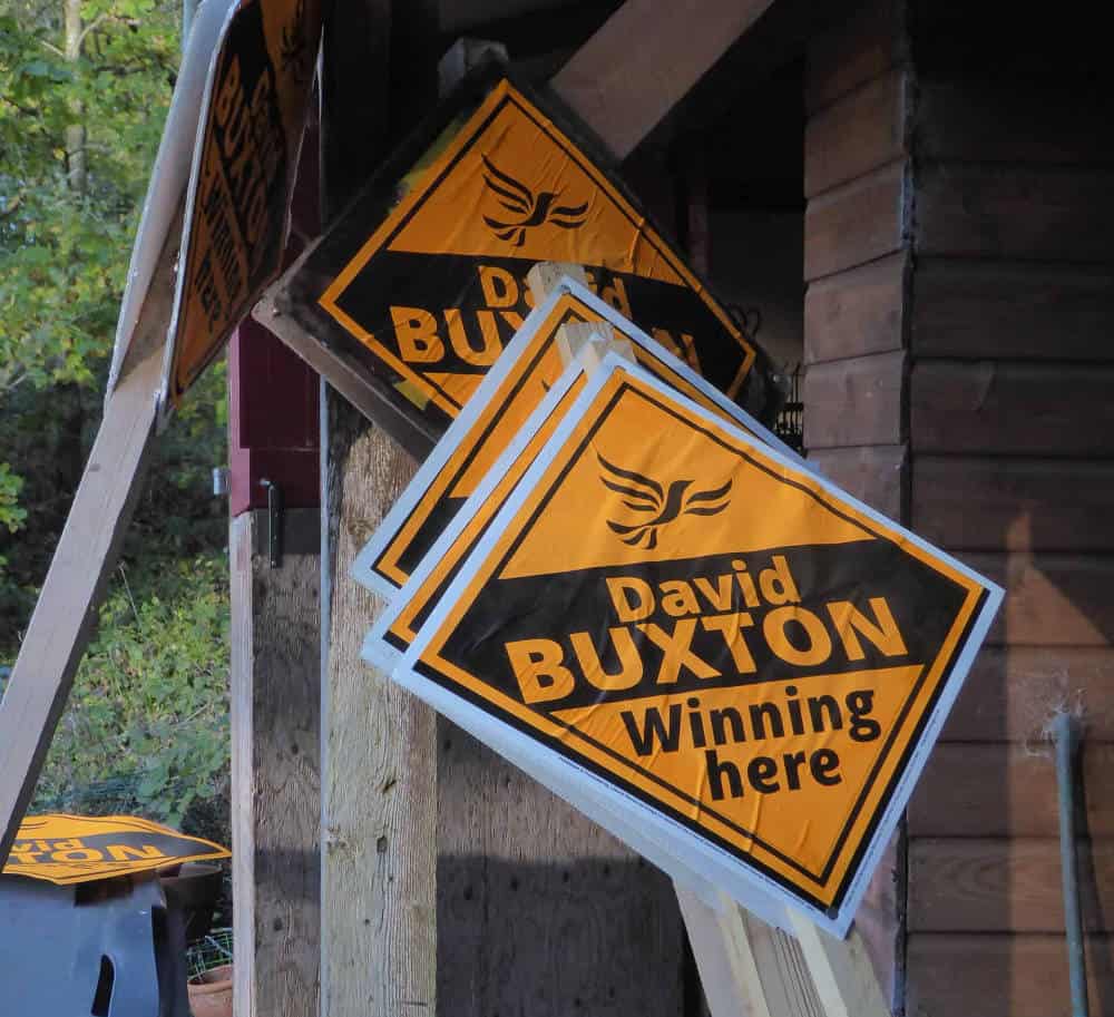 Damaged political campaign signs for "david buxton" mounted on a wooden post, with a blurred background showing greenery and part of a building.