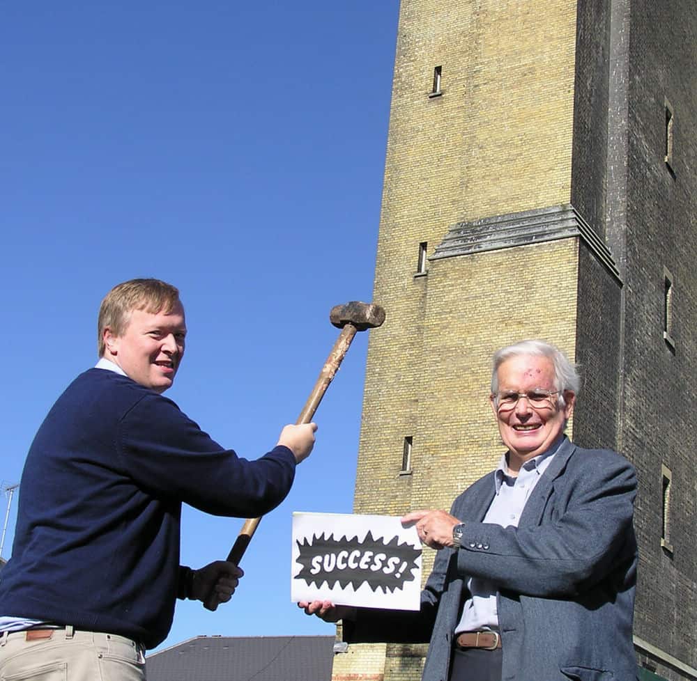Two men, one younger and one older, cheerfully holding a sledgehammer and a sign that says "success" respectively, standing outdoors next to a brick building.