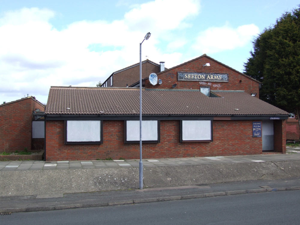 A closed pub, named "sefton arms," with boarded-up windows and a security camera, under a cloudy sky.