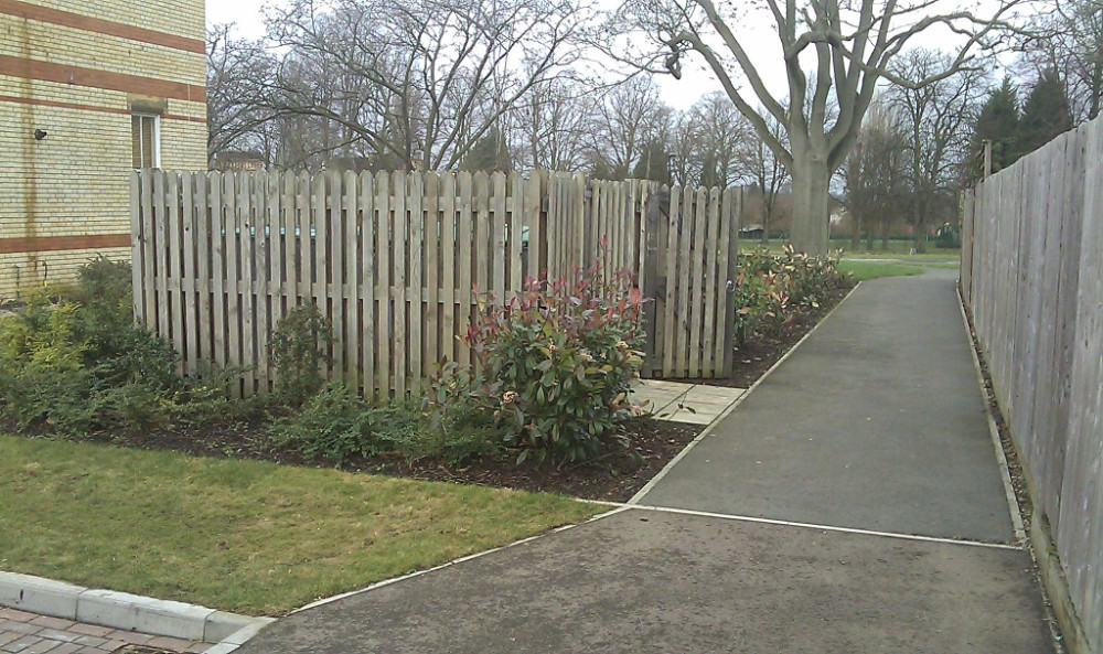 A wooden fence with a garden bed containing shrubs and flowers, next to a concrete pathway leading to a park.