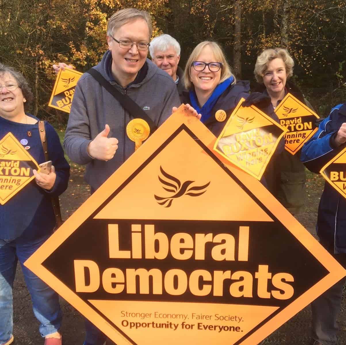 Group of smiling people holding a large liberal democrats sign and campaign placards in an outdoor setting.
