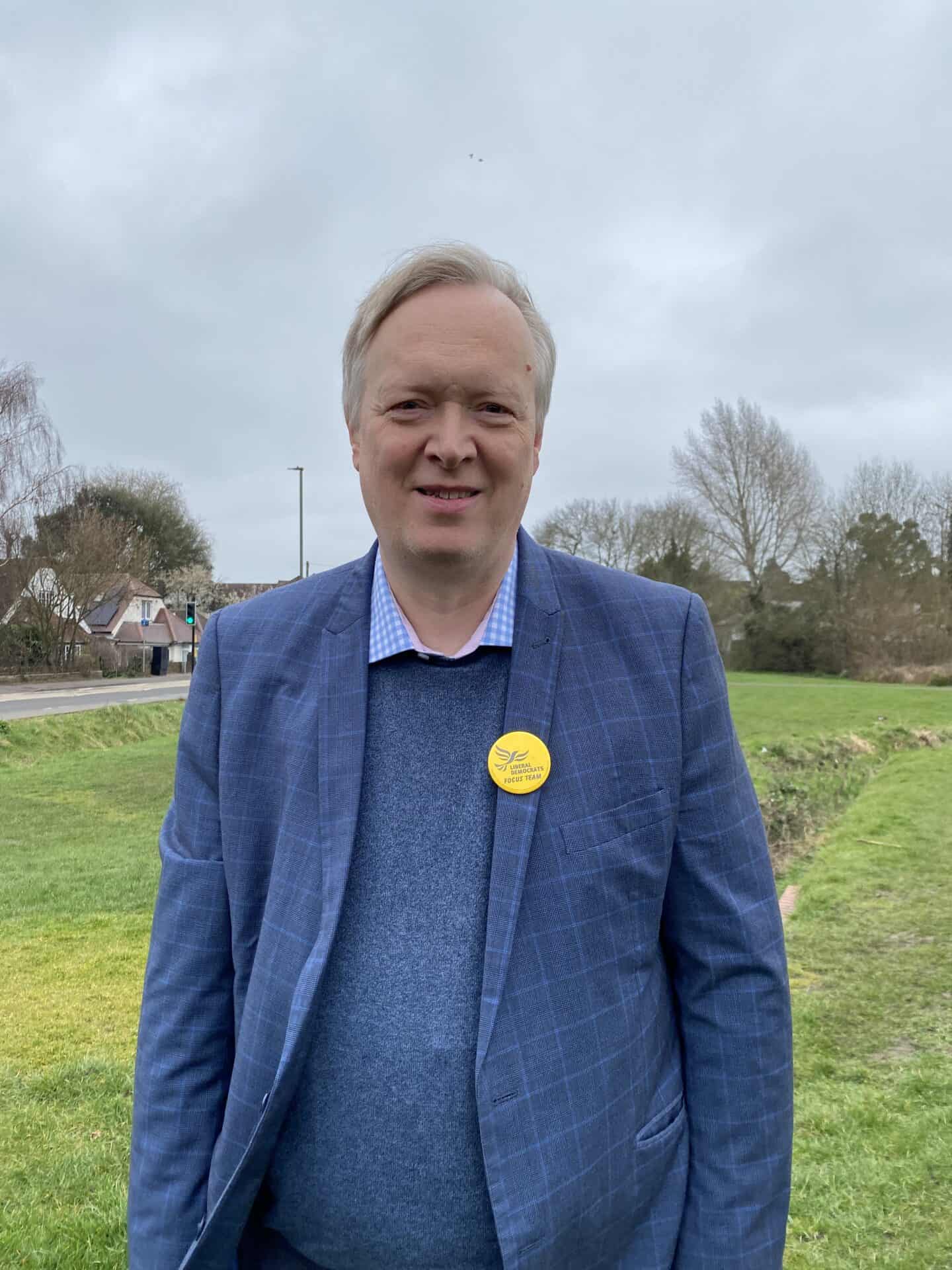 A middle-aged man with short blonde hair and a blue blazer over a pink shirt, wearing a yellow badge, stands outdoors with a grassy area and trees in the background.