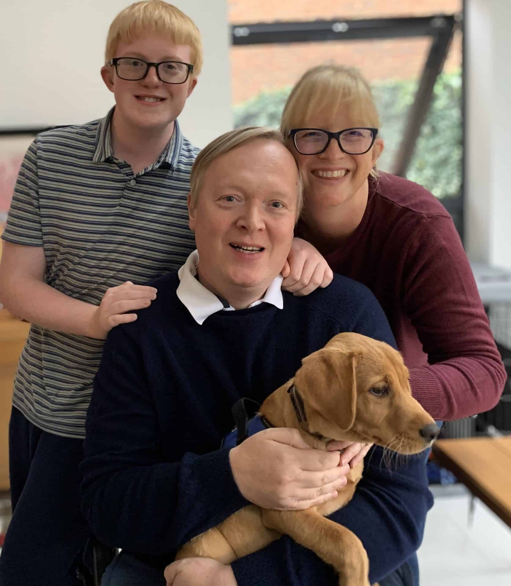 A family of three smiling with a young boy standing behind a seated couple holding a small dog inside a room.