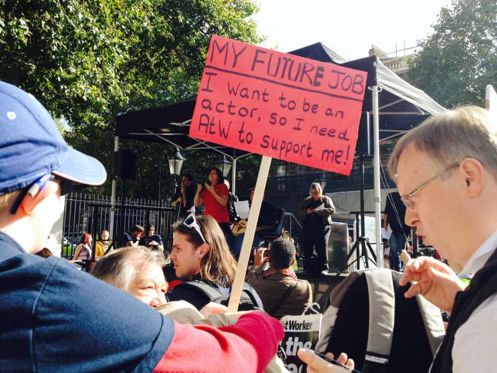 Person at a protest holding a sign that reads "my future job: i want to be an actor, so i need atw to support me!" with others around in a sunny outdoor setting.