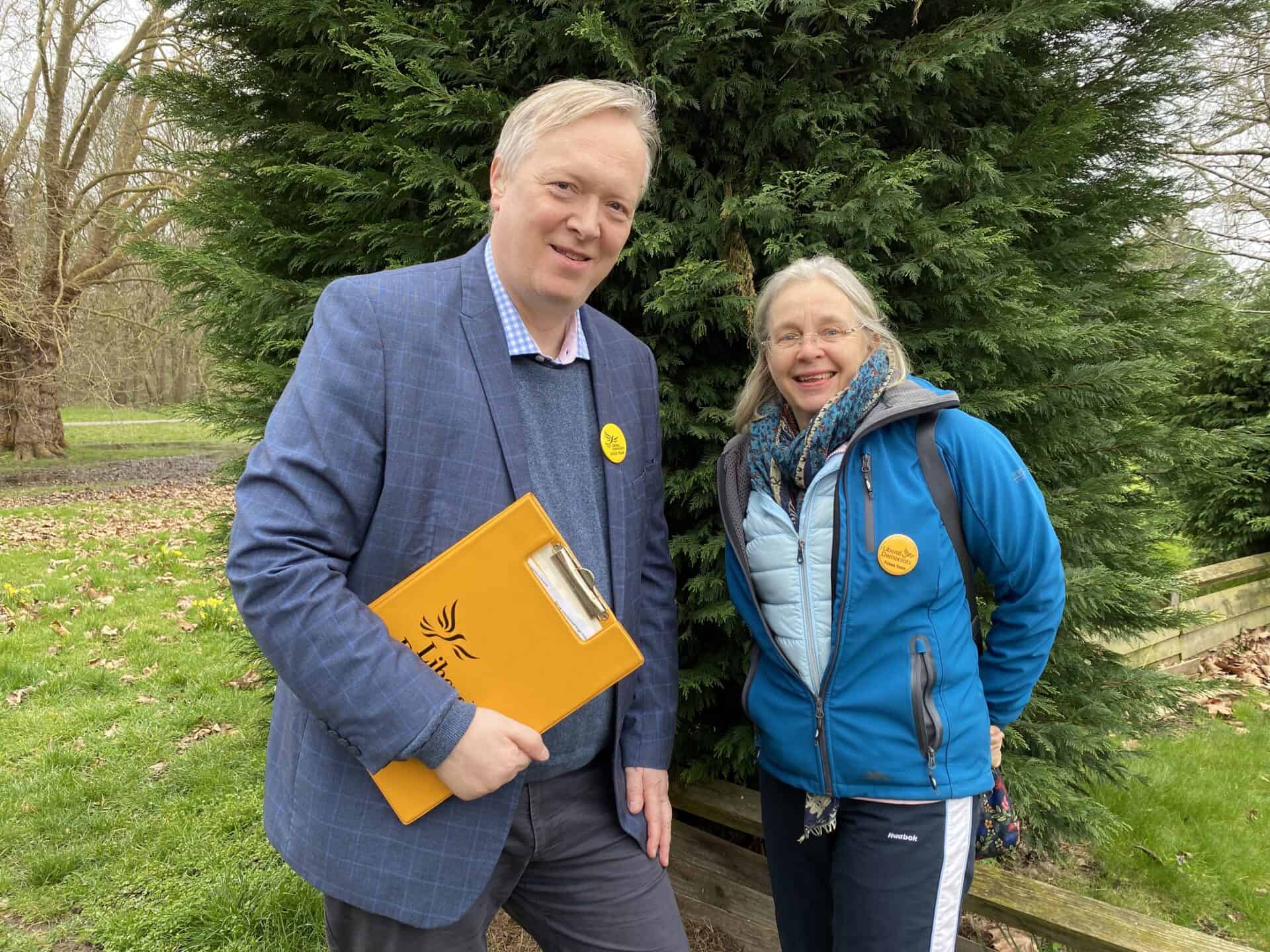 A man in a suit holding a clipboard and a woman in a blue jacket smile outdoors near trees, both wearing circular yellow badges.